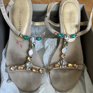Beige sandaler med stenar från Tamaris - Eleganta beige sandaler från Tamaris dekorerade med färgglada stenar i blått, vitt och guld. Sandalerna har en smal remdesign och är perfekta för sommarens varma dagar.
