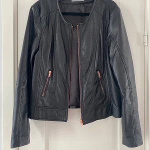 Svart skinnjacka med dragkedjor strl M - Snygg svart skinnjacka med coola dragkedjor i kopparfärg. Jackan har en stilren design med perforerade detaljer och långa ärmar. Perfekt för en edgy look! Svart kortare skinnjacka i strl M från Soaked in Luxury, modell Laia jack.  Endast använd vid ett par tillfällen och är i nyskick.  Mått Över bröstet: ca 51 cm Längd från axeln och ner: ca 57 cm  100% fårskinn jacka och 100% polyester foder  Kommer från ett djur och rökfritt hem.