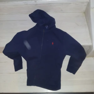 Mörkblå hoodie från Ralph Lauren - Säljer en snygg mörkblå hoodie från Ralph Lauren med dragkedja och det klassiska röda logotypbroderiet på bröstet. Perfekt för en avslappnad stil. Hoodien har långa ärmar och en bekväm passform.