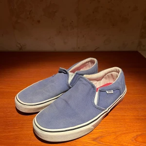 Blå slip-on sneakers från Vans - Säljer ett par klassiska blå slip-on sneakers från Vans. Skorna har en vit sula och är tillverkade i textilmaterial. Perfekta för en avslappnad stil.