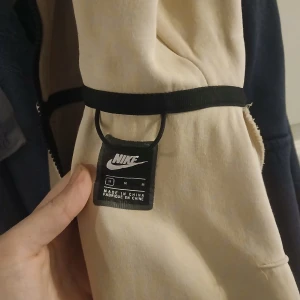 Beige och vit kofta från Nike - Säljer en snygg beige och vit kofta från Nike med svarta detaljer. Jackan har dragkedja och en stilren design. Perfekt för en sportig look.