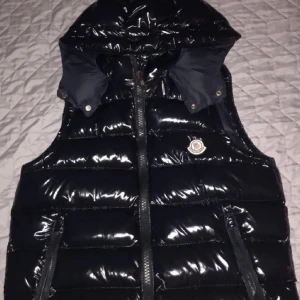 Svart dunväst från Moncler - Snygg svart dunväst från Moncler med glansig yta och avtagbar huva. Västen har en dragkedja framtill och två fickor med dragkedjor. Perfekt för kyligare dagar när du vill hålla stilen. Moncler-logga på bröstet ger en exklusiv touch har använt den två gånger 