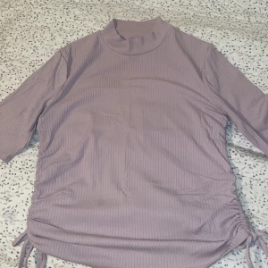 Lila ribbad topp från Lindex - Säljer en snygg lila ribbad topp från Lindex med trekvartsärm och knytning i sidorna.Toppen har en hög krage som ger en elegant touch. 💜