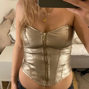 Guldig korsett med dragkedja - Snygg guldig korsett i glansigt material med dragkedja framtill. Den är i jättebra skick 