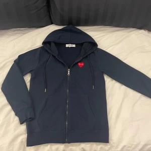 Mörkblå hoodie från Comme des Garçons - Säljer en snygg mörkblå hoodie från Comme des Garçons med det ikoniska röda hjärtat på bröstet. Hoodien har dragkedja och en skön passform med långa ärmar. Perfekt för en stilren och avslappnad look.