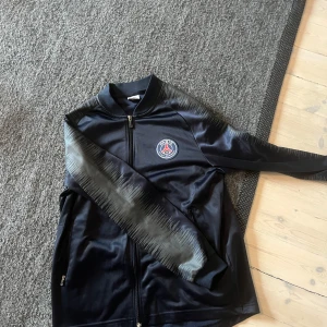 Blå PSG träningsjacka  - Snygg svart träningsjacka  med PSG-logga på bröstet. Jackan har en dragkedja framtill och en cool grå design på armarna. Perfekt för sportiga aktiviteter eller som en stilren vardagsjacka.