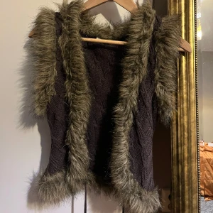 Brun stickad väst med fuskpäls - Säljer en brun stickad väst med mysig fuskpäls längs kanterna. Västen har en fin kabelstickad design och knytband med tofsar framtill. Perfekt för att hålla sig varm och stilren under kyliga dagar.  Skulle säga storlek xs-S