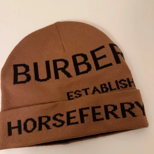 Beige mössa från Burberry - Snygg beige mössa från Burberry med svart text. Perfekt för att hålla värmen med stil. Mössan har en uppvikt kant och är tillverkad i ett mjukt material.