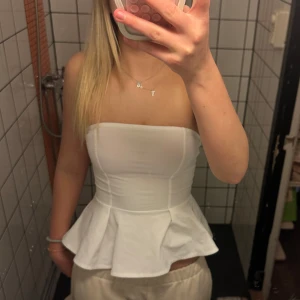 Vit volang topp från H&M - Säljer en stilren vit axelbandslös volang topp från H&M. Toppen har en peplumdesign som ger en fin siluett och är perfekt för att klä upp en outfit. 