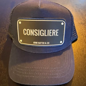 Blå keps från John Hatter & Co - Snygg blå keps med texten 'Consigliere' på framsidan. Kepsen har ett justerbart spänne bak och är tillverkad i en blandning av bomull och polyester. Perfekt för en avslappnad stil.