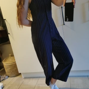 Randig jumpsuit med knappar - Snygg mörkblå Jump suit från Monki med vita ränder och knappar framtill. Den har smala axelband och en rak passform som ger en stilren look. Perfekt för en avslappnad men ändå chic stil. Axelbanden kan ställas in och göras längre eller kortare. Längd på hela jumpsuiten: 123cm. Ursäkta min skitiga spegel. 