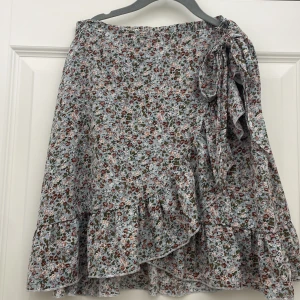 Blommig omlottkjol med volang - Charmig blommig omlottkjol i ljusblått med volangdetaljer. Kjolen har ett knytband i midjan och ett vackert blommönster i rosa och grönt. Perfekt för en somrig look!