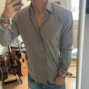 Burberry skjorta - Snygg grå skjorta från Burberry Brit med klassiska rutor på insidan av ärmsluten. Skjortan har långa ärmar och knappar framtill. Perfekt för en stilren look.