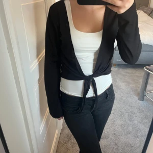 Svart bolerotopp från Happy Holly - Säljer en stilren svart bolerotopp från Happy Holly. Toppen har långa ärmar och ett knytband framtill som ger en snygg detalj. Perfekt att bära över en topp eller klänning för en extra touch. Passar bra till både vardag och fest.