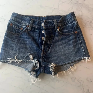 Blå jeansshorts från Levi's - Snygga blå jeansshorts från Levi's med fransiga kanter och knappar framtill. Perfekta för en avslappnad stil. Klassisk femficksdesign och högmidjade för en bekväm passform.