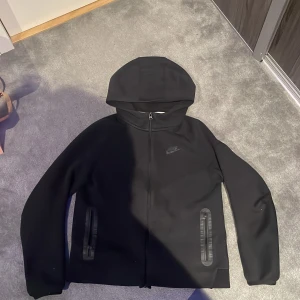 Svart hoodie från Nike - Säljer en svart Nike tech hoodie. Den är lite uppskrapad på armen som du kan se på bild 3, men det är inget som syns när du har på dig den. Den sitter bra och den ser bra ut både uppdragen och neddragen 