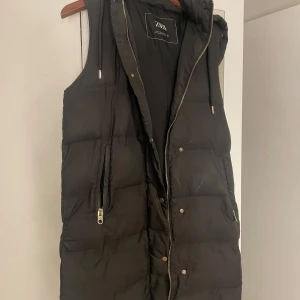 Svart dunväst från Zara - Snygg svart dunväst från Zara med dragkedja och knappar. Perfekt för kyliga dagar med sin quiltade design och huva. Västen har en stilren look och passar till många olika outfits.