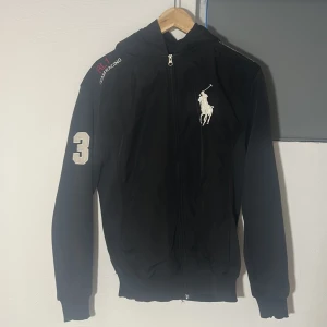 Svart cardigan från Ralph Lauren - Snygg svart cardigan från Ralph Lauren med dragkedja och huva. Den har ett stort vitt Polo-logotyptryck på bröstet och texten 'Ralph Lauren' på ryggen. Detaljer som 'Technical Crew' och 'All Garage Access' ger en unik touch. Perfekt för en stilren look. Skriv för fler bilder och passform etc:)