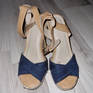 Beige och blå kilklackssandaler - Snygga kilklackssandaler med beige och blå detaljer. Sandalerna har en flätad kilklack och justerbara remmar runt ankeln. Perfekta för sommarens alla tillfällen.