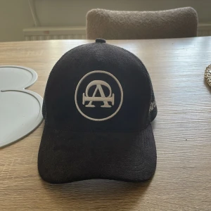 Keps - Snygg svart keps med vit broderad logga framtill och texten 'All Cap One' på sidan. Kepsen har ett nätmaterial baktill för bättre ventilation och en justerbar rem för perfekt passform.