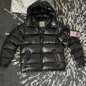 Svart pufferjacka från Moncler - Snygg svart pufferjacka från Moncler med glansig finish och dragkedja framtill. Jackan har en huva och Moncler-logga på bröstet. Perfekt för kyliga dagar när du vill hålla stilen.
