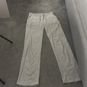 Grå mjukisbyxor från Juicy Couture - Säljer ett par grå mjukisbyxor från Juicy Couture. Byxorna har en bekväm passform med elastisk midja och snörning. De har två framfickor och två bakfickor med knappar. Perfekta för en avslappnad stil.