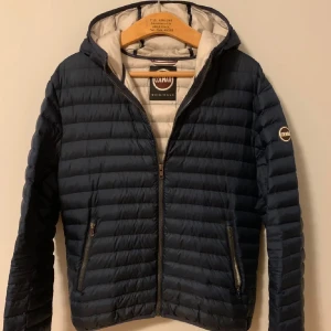 Mörkblå pufferjacka från Colmar - Säljer en snygg mörkblå pufferjacka från Colmar. Jackan har en dragkedja framtill och en huva för extra skydd. Den är perfekt för kyligare dagar och har en stilren design med Colmar-logga på ärmen.