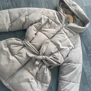 Beige pufferjacka från Nelly - Säljer en snygg beige pufferjacka från Nelly med huva och dragkedja. Jackan har ett bälte i midjan för en justerbar passform och quiltad design för extra värme. St 32 men är lite overaize så den är mer som st 34. Använd 2 ggr. 