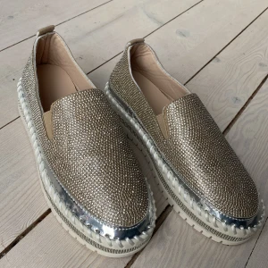 Glittriga loafers med silverdetaljer - Glitterloafers med silverdetaljer och vit rejäl sula. Bekväma men ändå 100 procent redo för fest, eller till och med under brudklänningen. För små för mig som är en bred 38. Kan funka fint för dig med storlek 37. Endast provade en gång inomhus. Sömmen har vikt in sig lite på ett ställe på hälen, se sista bilden. 