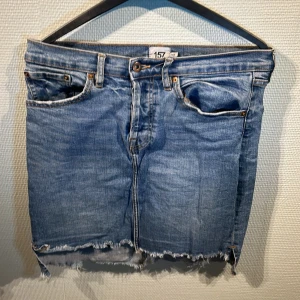 Blå jeanskjol  - Snygg blå jeanskjol från lager 157 med rå kant och klassisk femficksdesign. Kjolen har en knappgylf framtill och är perfekt för en avslappnad stil. Den är i en klassisk blå denimfärg och har en kort längd.
