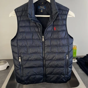 Mörkblå dunväst från Polo Ralph Lauren - Säljer en snygg mörkblå dunväst från Polo Ralph Lauren med dragkedja och broderad logga i rött på bröstet. Västen har en quiltad design och två praktiska fickor med dragkedja. Perfekt för dig med en stilren look. Passar dig som är mellan 155-165 cm lång.