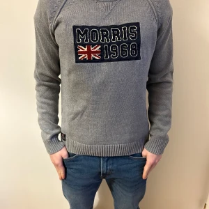 Grå stickad tröja från Morris - Säljer en stilren grå stickad tröja från Morris med ett broderat emblem på bröstet som visar 'Morris 1968' och en brittisk flagga. Tröjan har långa ärmar och ribbade muddar vid ärmslut och nederkant. Perfekt för en klassisk look.