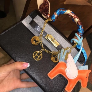 Svart handväska med kedjedetaljer - Snygg svart handväska med guldfärgad kedja och hängande berlocker. Väskan har ett färgglatt handtag med mönstrad scarf och en orange hästberlock. Perfekt för att ge en touch av stil till din outfit. Helt ny o oanvänd 