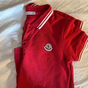 Röd pikétröja från Moncler - Säljer en snygg röd pikétröja från Moncler med klassisk krage och vita ränder. Tröjan har korta ärmar och Monclers logotyp broderad på bröstet. Perfekt för en stilren look!
