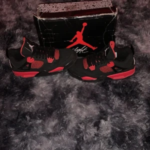 Jordan 4 red thunders - Snygga svarta och röda Nike Air Jordans med ikonisk design. Skorna har snörning och en vit Jumpman-logga på tungan. Perfekta för den som vill ha en stilren och sportig look.