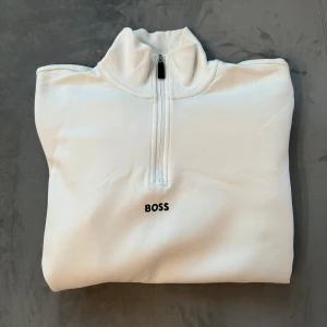 Vit tröja med dragkedja från Boss - Säljer en stilren vit tröja från Boss med halv dragkedja och broderad logga på bröstet. Tröjan har långa ärmar med ett mönster av Boss-loggor längs ärmarna. Perfekt för en avslappnad och trendig look. Vid frågor är det bara att höra av er.
