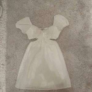 Vit klänning med puffärmar - Charmig vit klänning med puffiga kortärmar och ett sött blommigt mönster. Klänningen har en knytning framtill och små utskärningar vid midjan för en extra touch. Perfekt för studenten eller ett annat somrigt tillfälle🌸