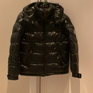 Svart dunjacka från Moncler - Snygg svart dunjacka från Moncler med glansig finish och huva. Jackan har dragkedja och långa ärmar, perfekt för kyliga dagar. Märkeslogga på ärmen ger en exklusiv touch. Men den har varit använd många gånger men den är ändå i fin skick