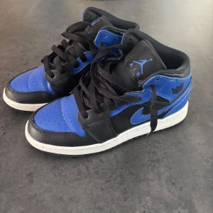 Nike Air Jordan 1 Mid i svart och blå - Snygga Nike Air Jordan 1 Mid sneakers i svart och blått. Skorna har en klassisk design med snörning och en bekväm sula. Box ingår.