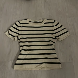 Randig kortärmad tröja från Zara - Säljer en stilren randig tröja från Zara i svart och beige. Tröjan har korta ärmar och en rund halsringning. Perfekt för en avslappnad look.