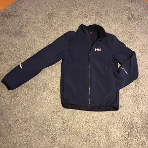 Mörkblå jacka från Helly Hansen - Säljer en stilren mörkblå jacka från Helly Hansen med dragkedja framtill och logga på bröstet. Jackan har reflekterande detaljer och är perfekt för kyligare dagar. Den är tillverkad i ett slitstarkt material som håller dig varm och bekväm. Jackan är i storlek 164/14år!