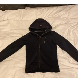 Svart huvtröja från Sail Racing - Säljer en svart huvtröja från Sail Racing med dragkedja framtill. Tröjan har en broderad logga på bröstet och en bekväm huva. Perfekt för kyliga dagar.