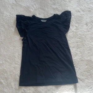 Svart topp med volangärmar från TIKI - Snygg svart topp från TIKI med volangärmar. Perfekt för en stilren look. Tillverkad i ett mjukt material som känns skönt mot huden.