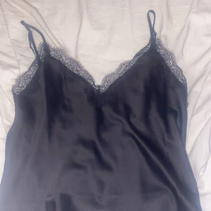 Svart linne med spetsdetaljer - Snyggt svart linne med spetsdetaljer längs halsringningen och tunna axelband. Perfekt för en elegant och stilren look.