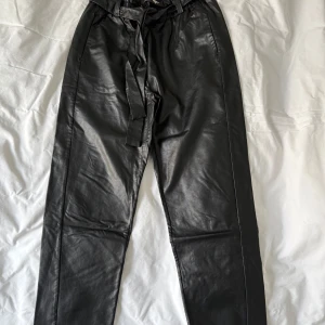 Svarta byxor i imiterad Skinn från House of Lola - Snygga svarta byxor i syntetmaterial från House of Lola. Byxorna har en hög midja med knytband och en stilren design. Perfekta för en trendig look. Byxorna sitter lite lösare och är supermjuka och sköna. Lite tunnare och med croppad längd. Jag är 163 cm lång.