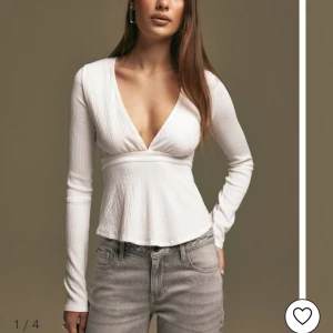 Vit ribbad topp med knyt - INTRESSEKOLL en superfin vit ribbad topp med v-ringning och långa ärmar. Toppen har en snygg knytning i ryggen som ger en extra detalj. Perfekt för både vardag och fest!