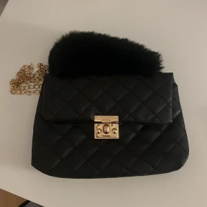 Svart quiltad handväska med guldkedja - Säljer en superfin svart quiltad handväska med en lyxig guldkedja och fluffig pälsdetalj på handtaget. Väskan har en elegant guldfärgad låsdetalj framtill. Perfekt för en utekväll eller fest! 🖤✨kan ha runt om sig 