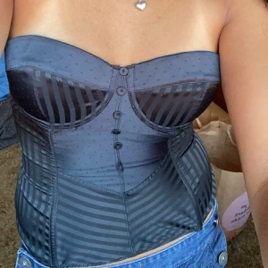 Svart korsett med randigt mönster - Säljer en snygg svart korsett med randigt mönster och prickiga detaljer. Den är axelbandslös och har knappar framtill. Perfekt för en festlig kväll eller en utekväll med vänner. 🌟