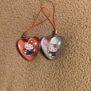 Hello Kitty hjärtformade hängen - Två supergulliga Hello Kitty-hängen i form av hjärtan. Det ena är rött med en söt Hello Kitty som har en rosett och det andra är blått med en Hello Kitty som håller en ballong. Perfekta som dekorationer eller att hänga på väskan!