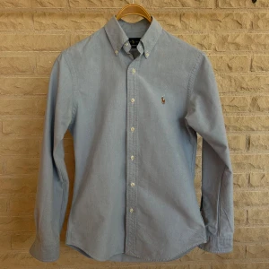 Ljusblå skjorta från Ralph Lauren - Säljer en stilren ljusblå skjorta från Ralph Lauren. Skjortan har en klassisk button-down krage och är dekorerad med det ikoniska broderade logotypen på bröstet. Den är långärmad och tillverkad i ett mjukt bomullsmaterial. Perfekt för både vardag och fest!